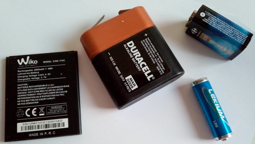 Fig. 1: Exemple de piles et batteries. De gauche à droite : Une batterie de Smartphone Li-ions, 4,2V, 2Ah, 7.4Wh. Puis trois types de piles alcalines, une pile de 4,5V. Une pile de 1,5V. Une pile de 9V, 0,55Ah